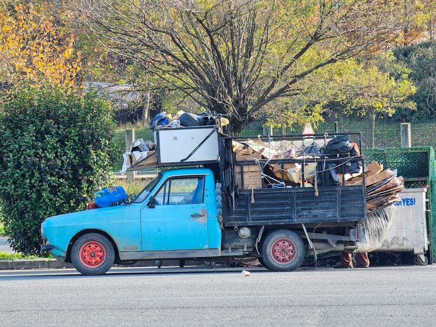 A sanitation worker dressed in a blue uniform with an orange high-visibility vest is discarding waste into the rear compartment of a large white rubbish collection truck parked alongside a narrow city